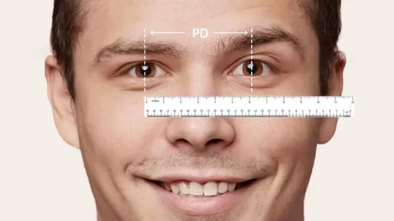 Close-up of a smiling man's face with a ruler graphic overlaid across his eyes, showing a dashed line labeled 'PD' connecting his pupils, illustrating the process of measuring pupillary distance.