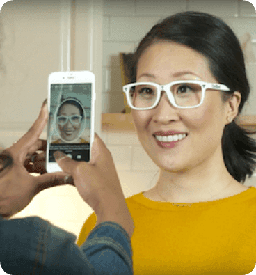 A smiling woman wearing white eyeglasses, taking a selfie or being photographed by someone holding a smartphone, with her face visible on the phone screen, illustrating a digital method for virtual try-on or PD measurement.